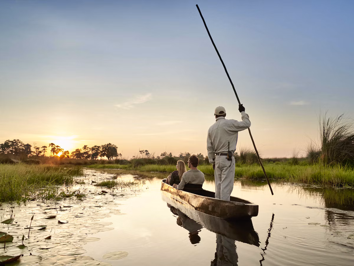 A guide poling a couple through a still Okavango channel by mokoro at sunset