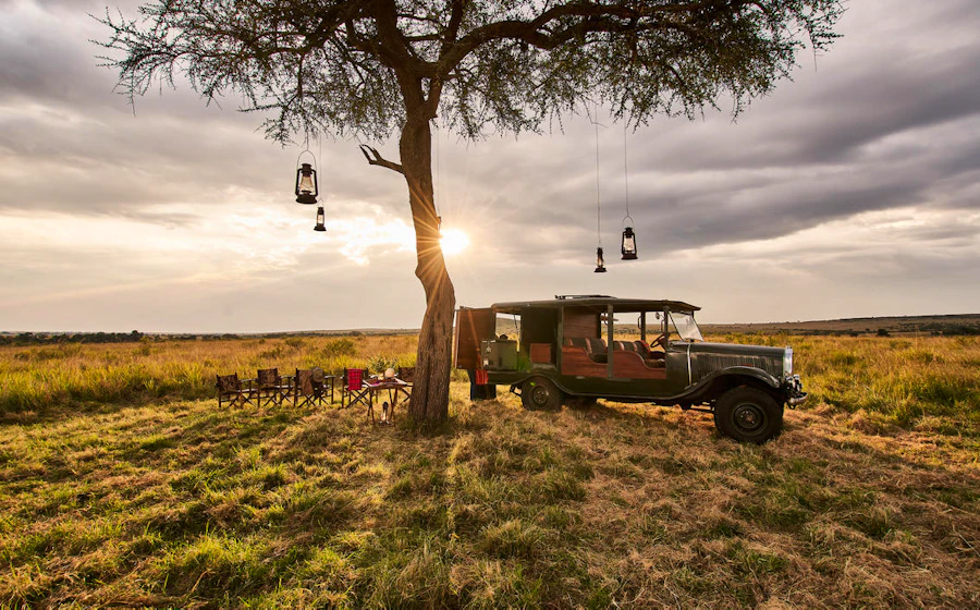 A private safari picnic beside a vehicle under a lantern-lit tree at sunset